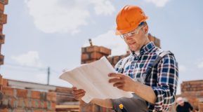 photo image of a builder in an orange hard hat, blue check shirt, looking at building plans